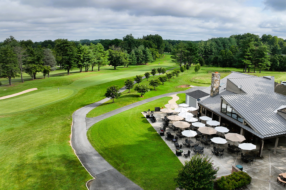Aerial view of the clubhouse at Enterprise Golf Course. There is a pathway surrounded by trees, and many outdoor tables with white and gray umbrellas on a patio next to the Clubhouse building.