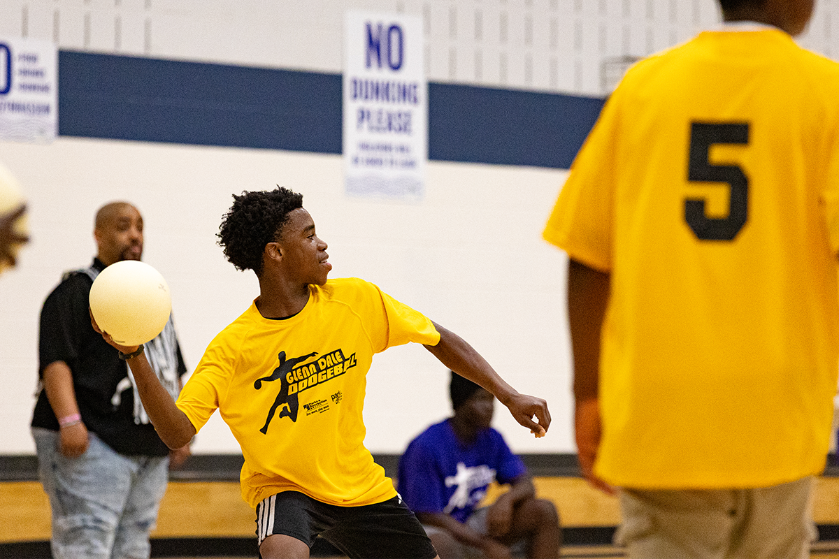 In a gymnasium, a young man in a yellow jersey leans back with a white ball in his right hand, preparing to throw it. The jersey reads Glenn Dale Dodgeball.