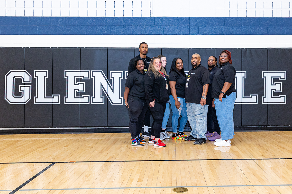 Eight adults in matching, black staff polo shirts pose in a gymnasium in front of a wall of mats with words GLENN DALE in white letters.