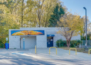 Exterior view of a single-story cinderblock building with a blue and gold sunrise mural on one wall.