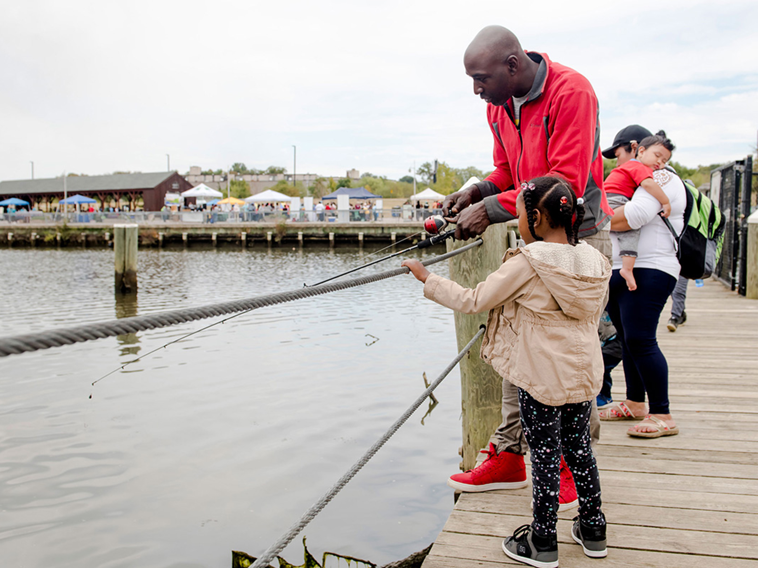 A man in a red jacket and red sneakers stands on a wooden pier or dock next to his daughter, holding a fishing pole. Behind him, a woman carries a sleeping toddler. In the distance, a row of event tents is set up between a picnic shelter and the water's edge.