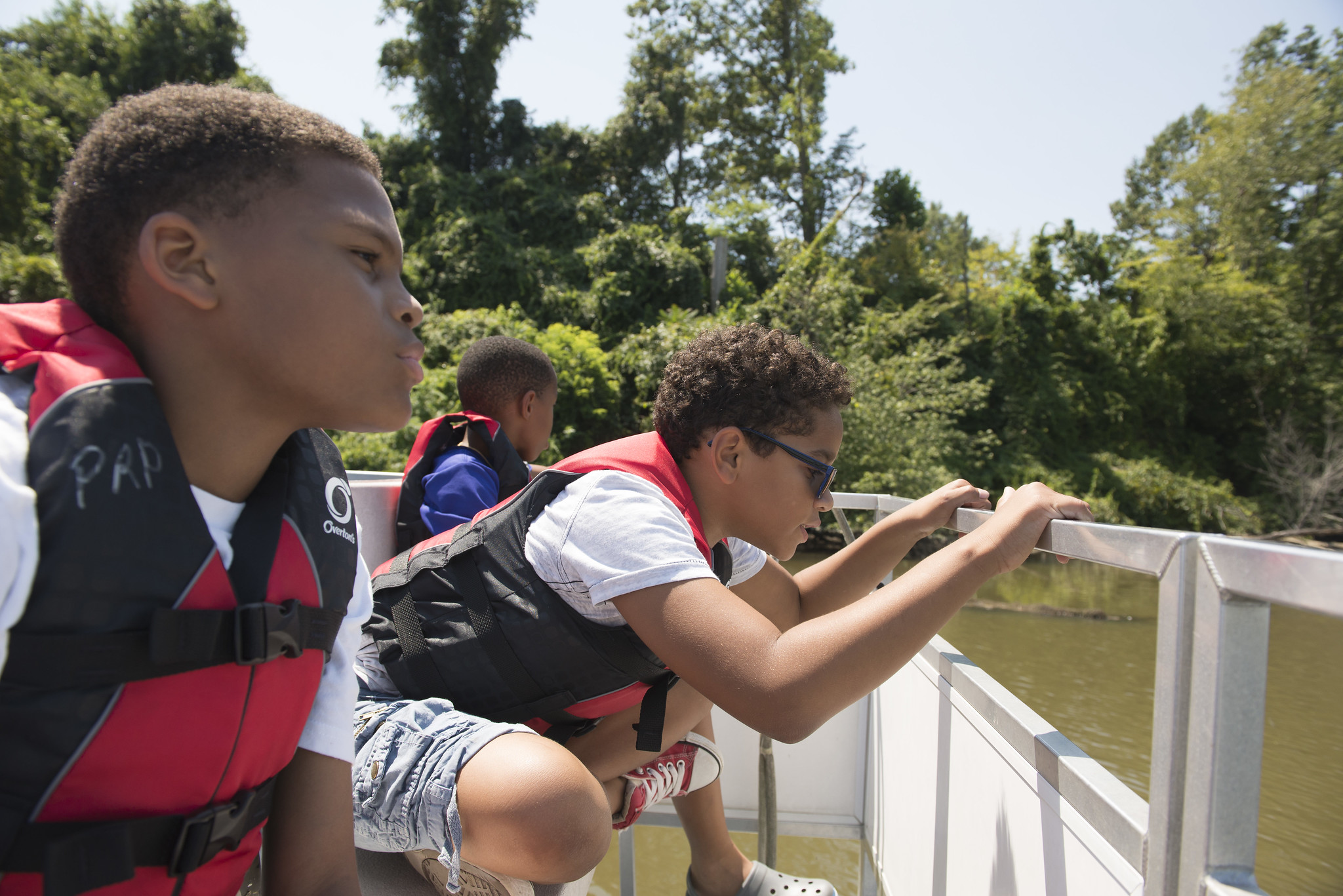Three youth wearing red life vests look over the railing of a boat at the greenish brownish water below.
