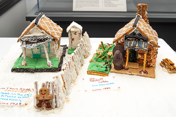 Two wonderful gingerbread log houses, with pretzel supported entryways. Both are adorned with black licorice decorations. There is a frosted pretzel stick fence between the two.
