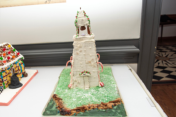 A tall white gingerbread lighthouse. It has candy cane supports, and a chocolate bell and light at the top.