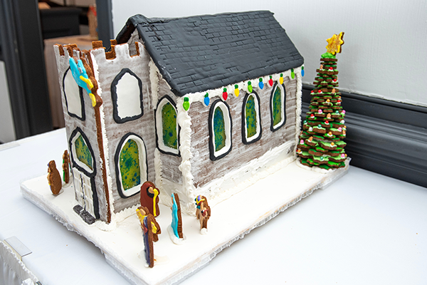 A gray gingerbread church with white icing mortar and molding to hold it together. It has a large front steeple, as well as lively green and blue sugar stained glass windows throughout. There is a gingerbread nativity scene out front, and a large gingerbread tree at the back corner of the building.
