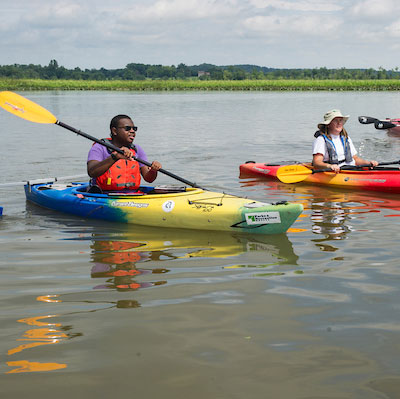 A man and a woman sit in plastic kayaks holding yellow and black paddles, in a body of water. The woman wears a sun hat and the man wears sunglasses.