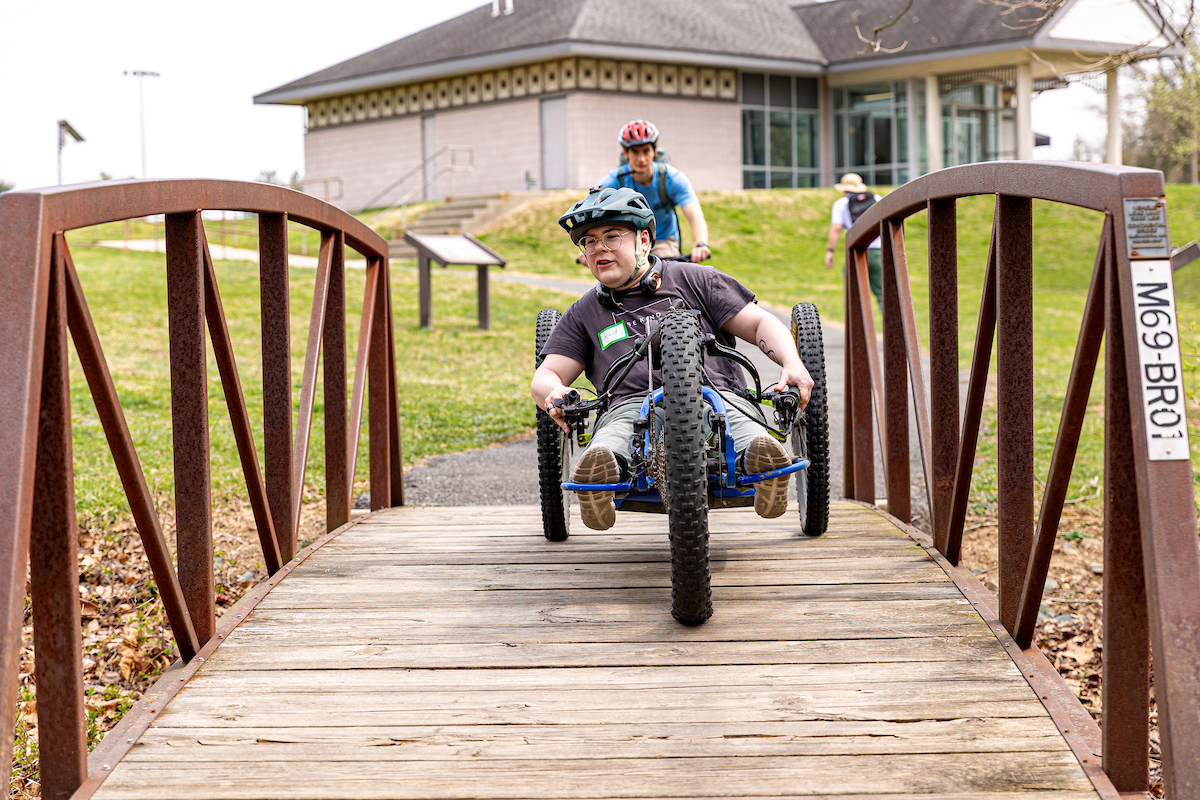 A person using an adaptive bike rides over a small wooden footbridge with metal trusses. Behind him is another cyclist on a paved path. There is a single-story beige building in the background.