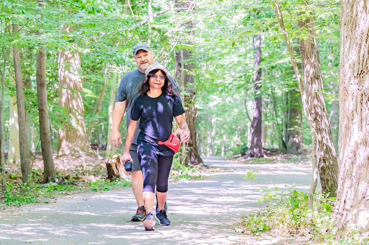 Two adults in fitness clothing walk along a wide, shady, paved path in a wooded area. Tall trees surround the path on both sides.