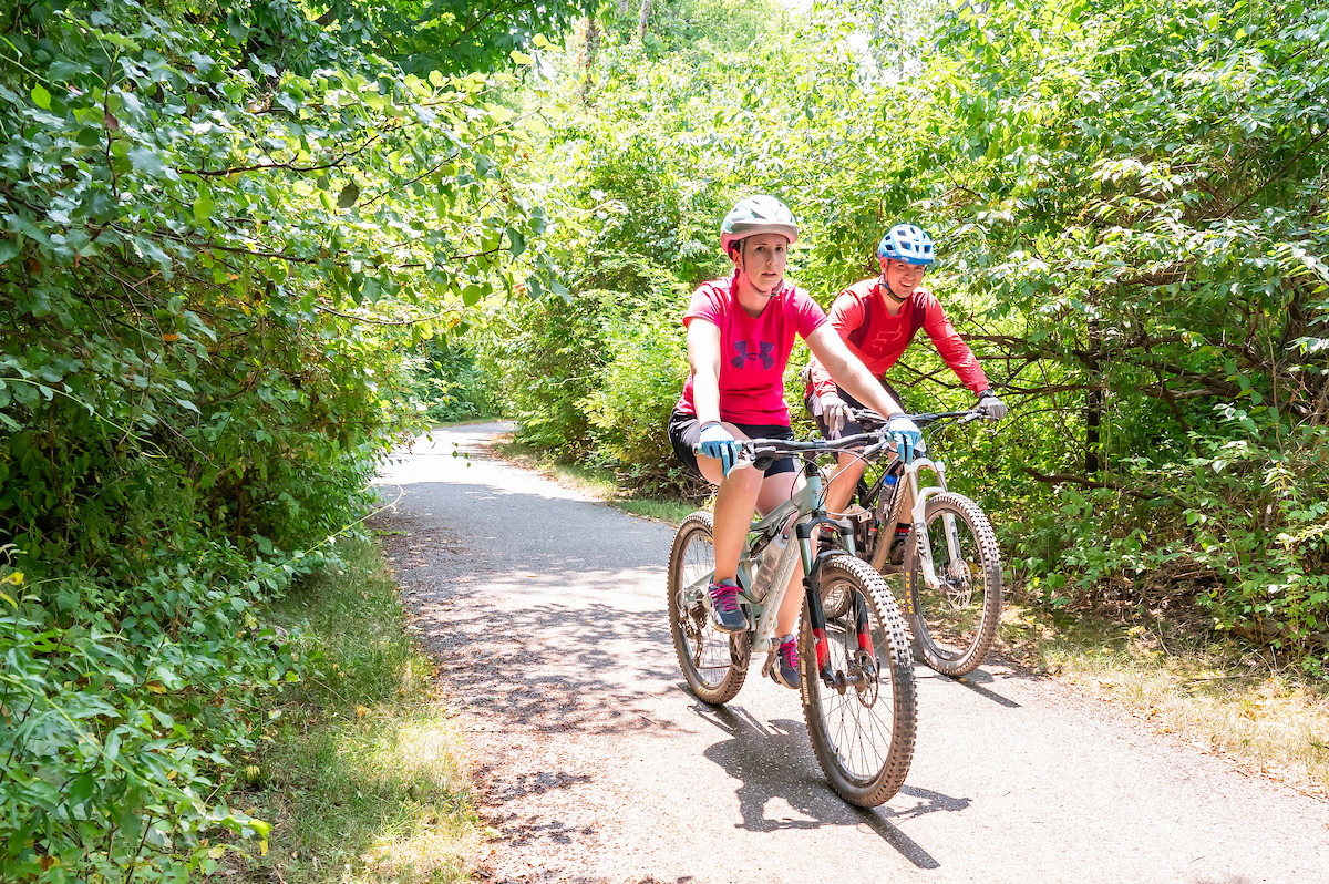 Two cyclists, both in red shirts, travel along a paved path. Leafy green foliage and trees surround the path on both sides. The path is partially shaded.