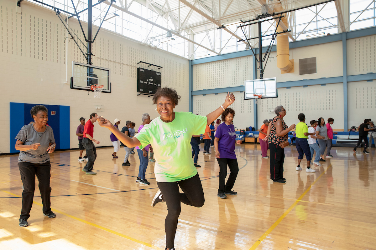 A class of seniors prepares to exercise in a gymnasium. Closest t the camera is a smiling woman in neon green with her arms raised.