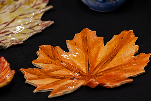 A colored and glazed ceramic leaf shaped plate on a black table. There are other similar ceramic leaves on the edges of the photo.