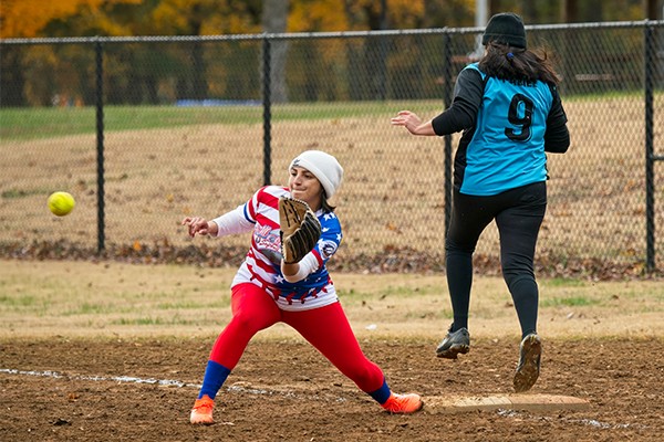 Two women play softball. One is leaning down, bracing to catch a ball and tag first base. The other is running through first base. The batter/runner is safe.