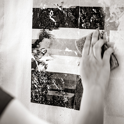 A black-and-white image of a hand using a sponge to transfer a photographic image onto the surface of a painting.