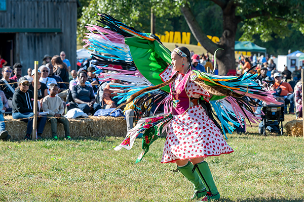 A performer in a colorful costume with a dramatically fringed cape does a traditional Native American dance outside on the grass. Behind a temporary fence, spectators sit on bales of hay near a barn, watching.