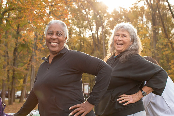Two women leaning on a picnic table, doing stretches preparing for a walk. There are many trees and leaves scattering the ground in the background