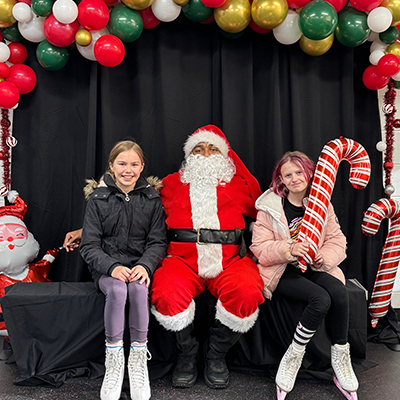Santa Claus sitting beside two girls surrounded by red, green, and white balloons. One girl is holding a giant candy cane.