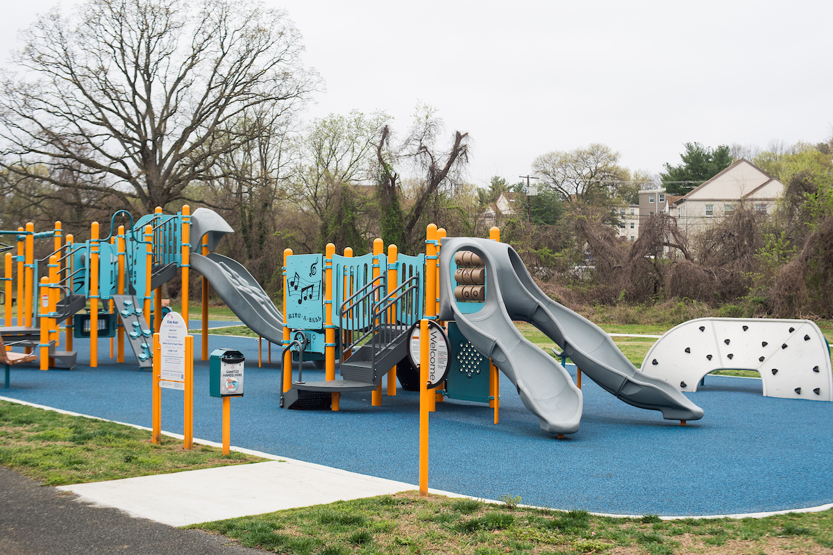 An outdoor playground with multiple play structures that feature slides, stairs, platforms, climbing walls, games, musical instruments, and spinning elements. The play equipment is light blue with yellow vertical posts.