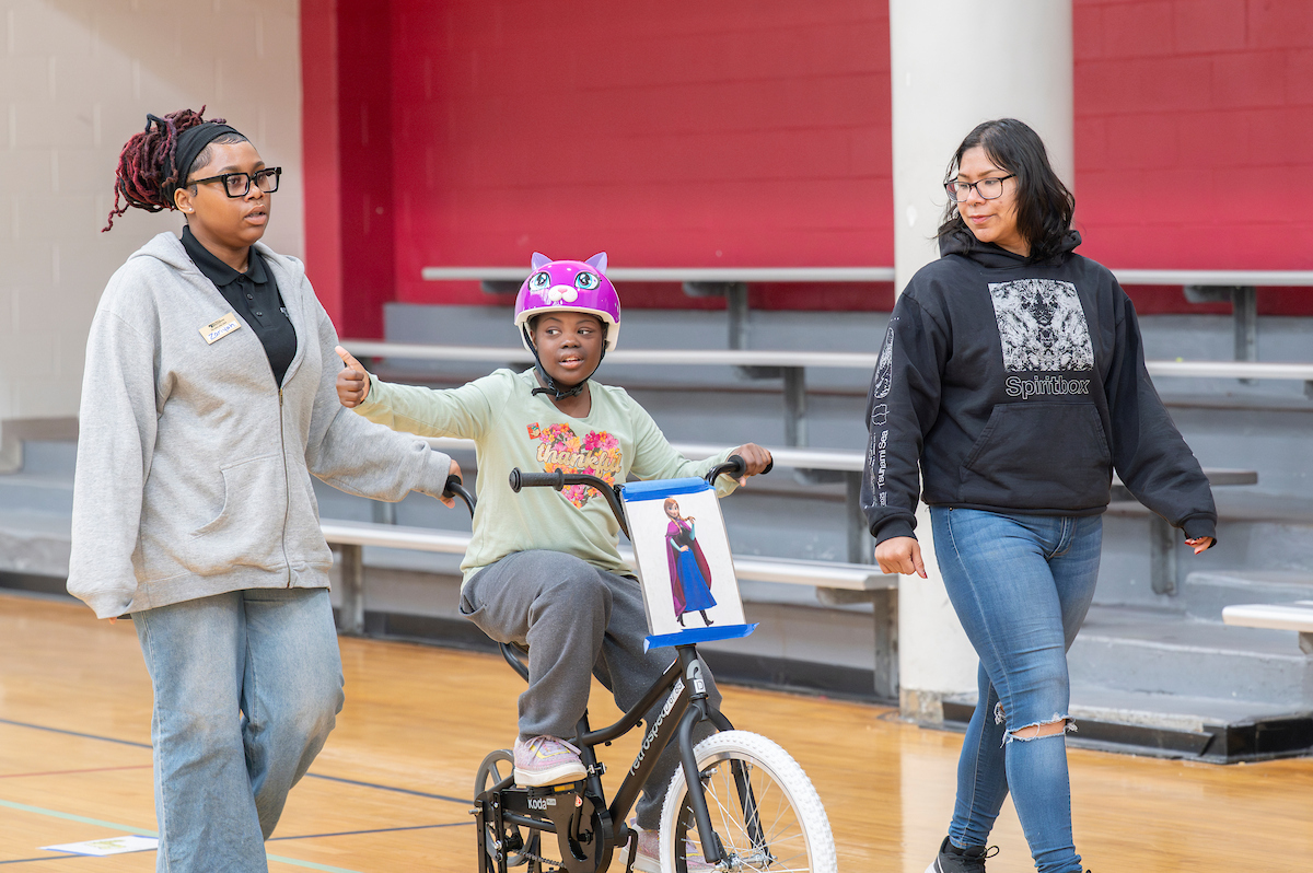 In a gymnasium, two adults walk alongside a youth on an adaptive bicycle. The youth wears a purple bike helmet that looks like a cat head, and gives a thumbs up with her right hand.
