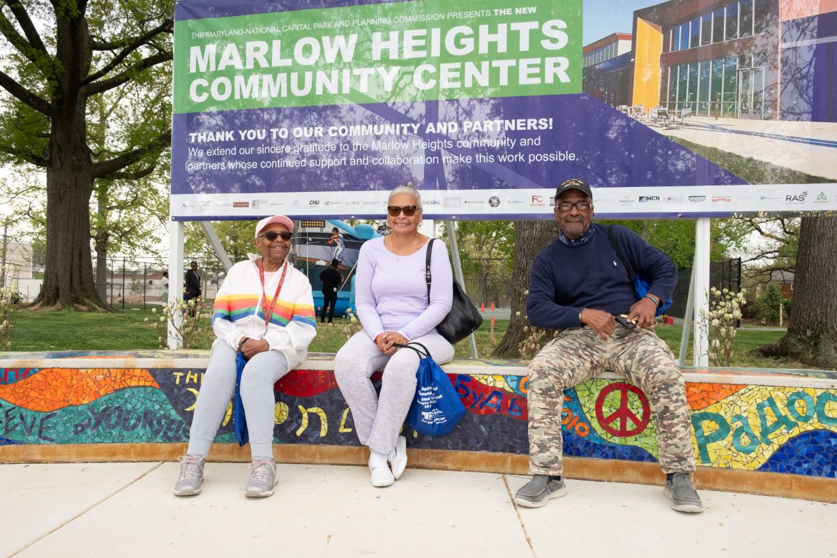 Three smiling adults sit on a long, curved, outdoor bench that features colorful mosaic artwork on the top and the sides. A large banner in the grass behind them reads MARLOW HEIGHTS COMMUNITY CENTER: THANK YOU TO OUR COMMUNITY AND PARTNERS!