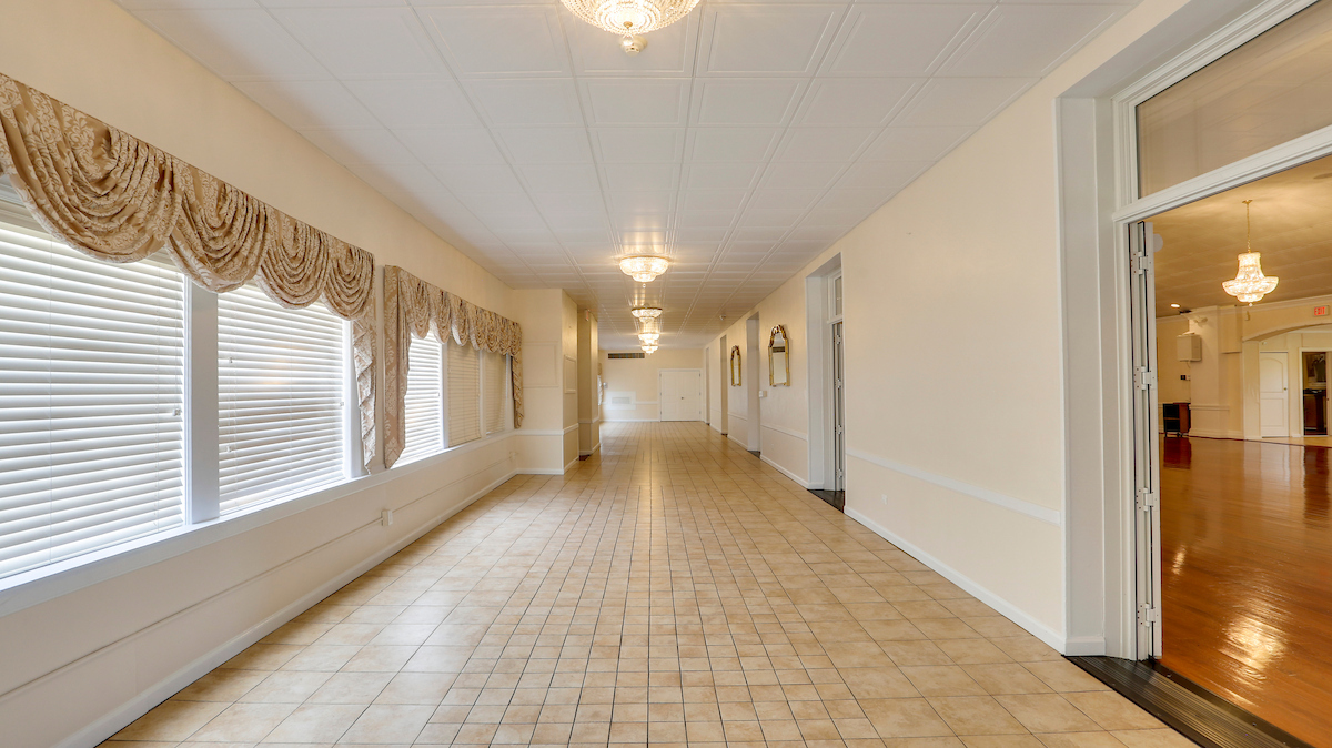 A long, tiled room situated off the ballroom, with windows along the left side and crystal light fixtures overhead. Multiple doorways along the right side lead into the ballroom through double doors.