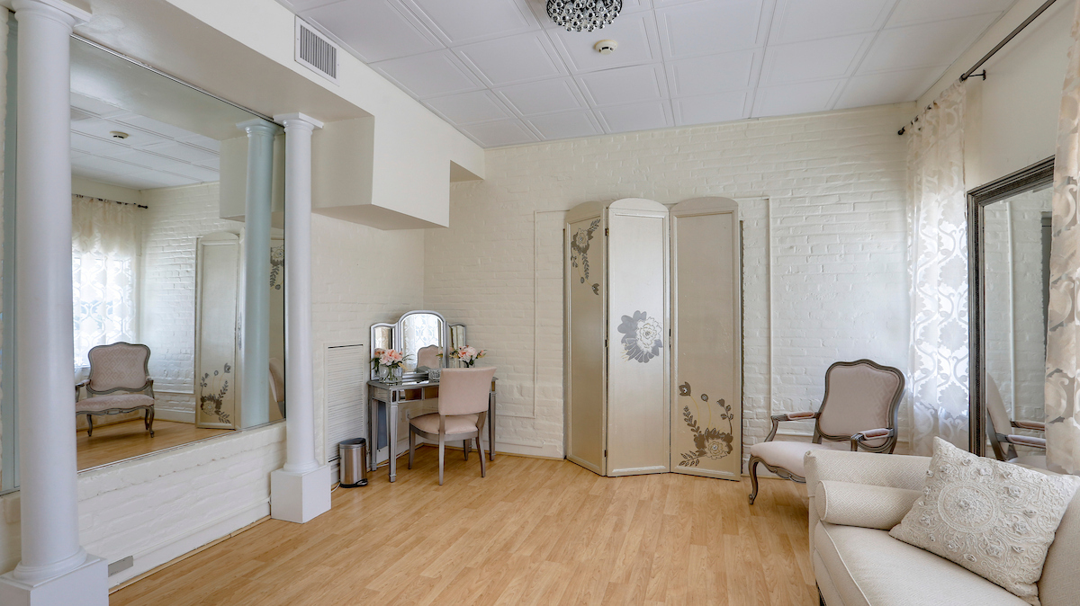 A small room with hardwood floors, large mirrors on multiple walls, an ivory sofa and armchair, a mirrored vanity table in one corner, and a privacy screen along one wall.