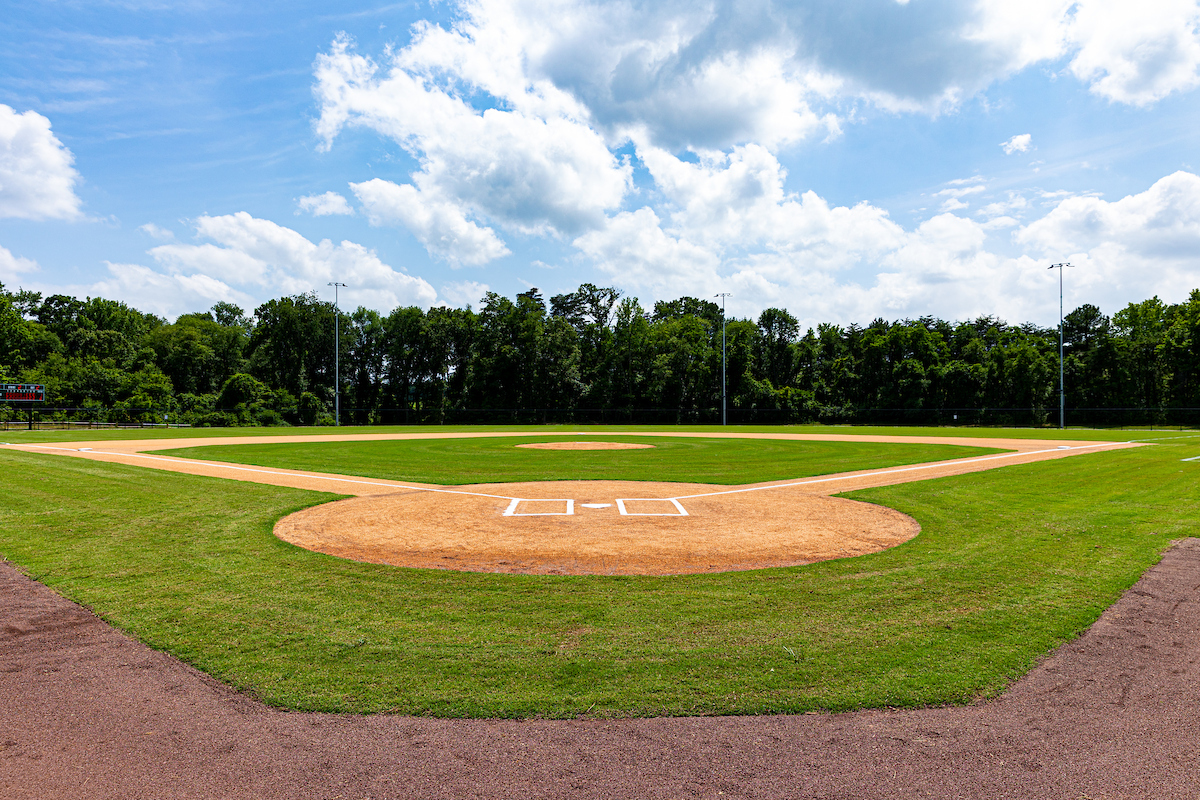 A large, brand new baseball diamond as seen from behind home plate, beneath a blue sky streaked with clouds. A scoreboard is installed in the outfield, along with several tall light poles for nighttime play.