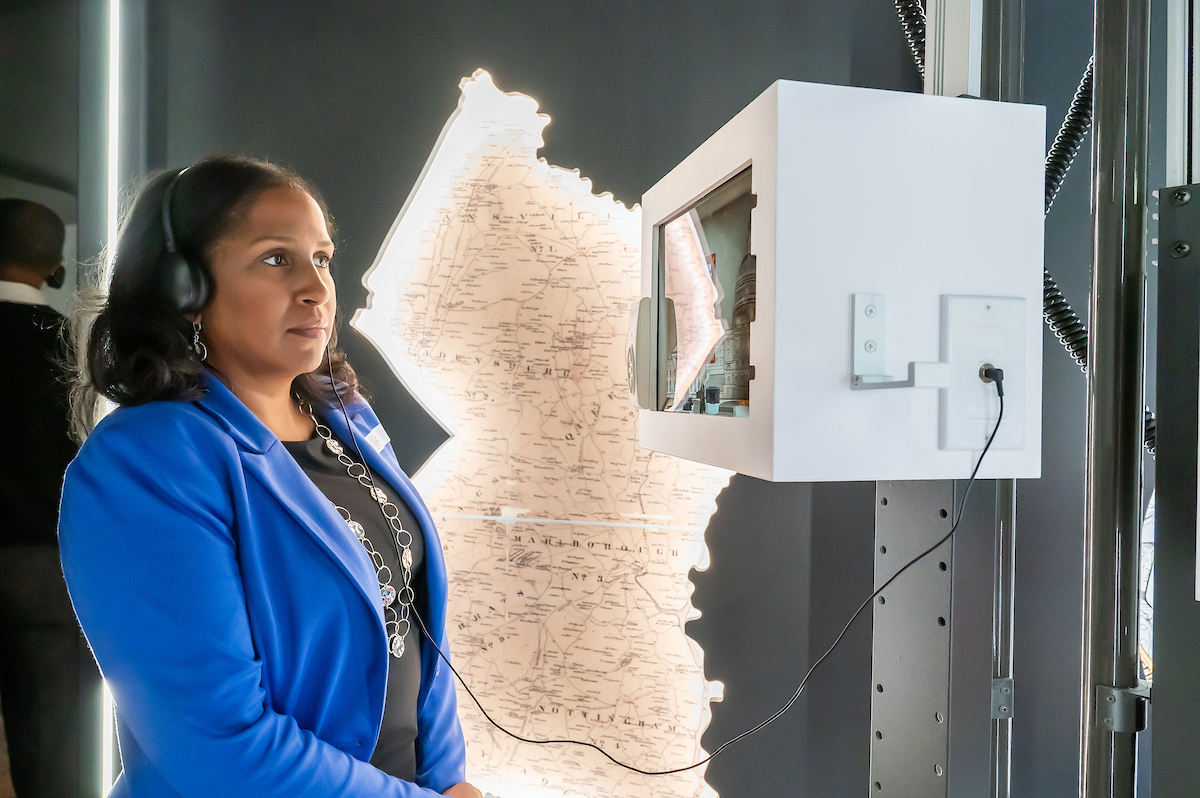 A woman in a blue suit jacket looks at a small video monitor while listening to headphones connected to it. Behind her is an illuminated map of Prince George's County.
