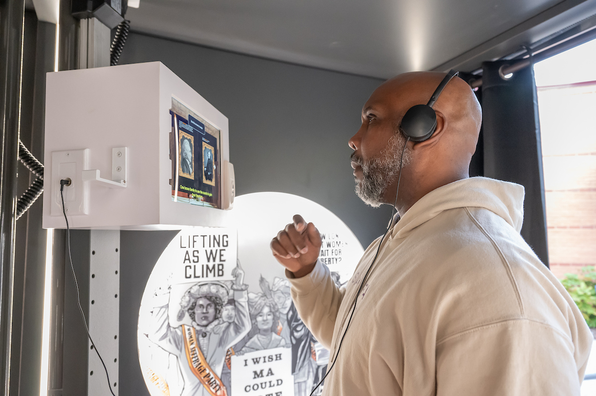 A man listens to headphones that are connected to museum exhibit in an interactive monitor. Behind him is a lighted panel with illustrations of protest marchers throughout history.