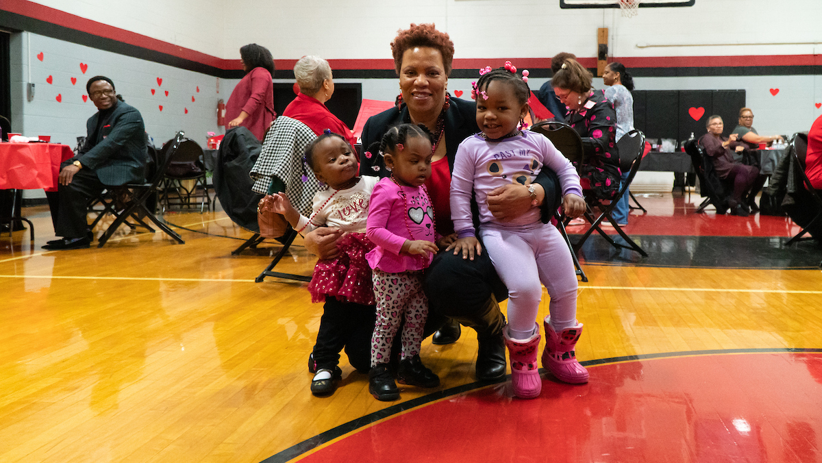 Inside a gymnasium decorated for Valentine's Day, a smiling woman crouches near the floor with her arms around three toddlers, all wearing hearts and Valentine-themed outfits. Behind them, people sit at decorated tables eating and visiting.