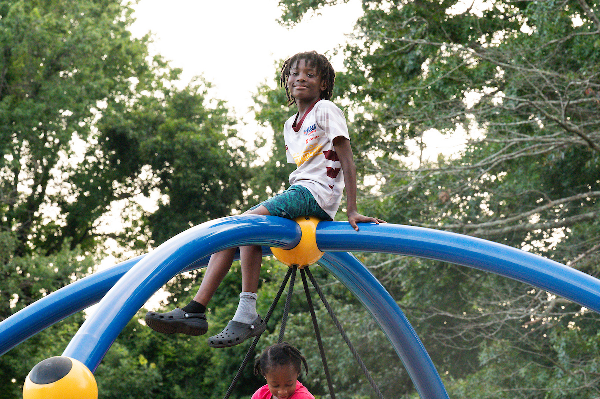 A smiling child sits atop a large swing support structure on an outdoor playground. Below him, a child in pink plays on a swing suspended from four ropes.