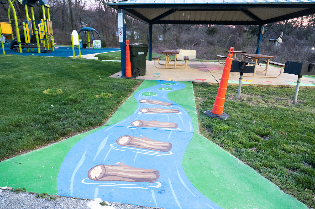 A short sidewalk that runs through grass to a picnic shelter is painted to resemble a river with logs floating in it. To the left of the picnic shelter is green and blue playground with several climbing structures.