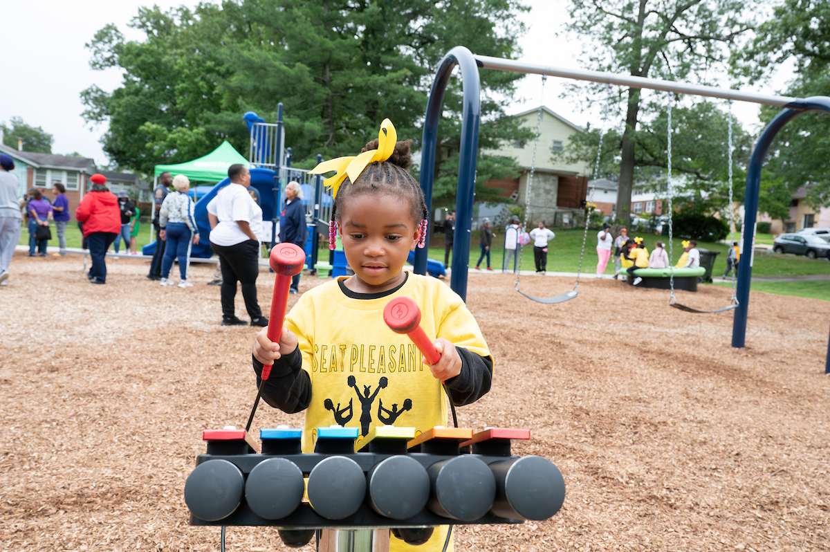 A child wearing a yellow shirt and a yellow ribbon in her hair uses red mallets to play a large xylophone at an outdoor playground. Behind her are swings slides, and other play structures.