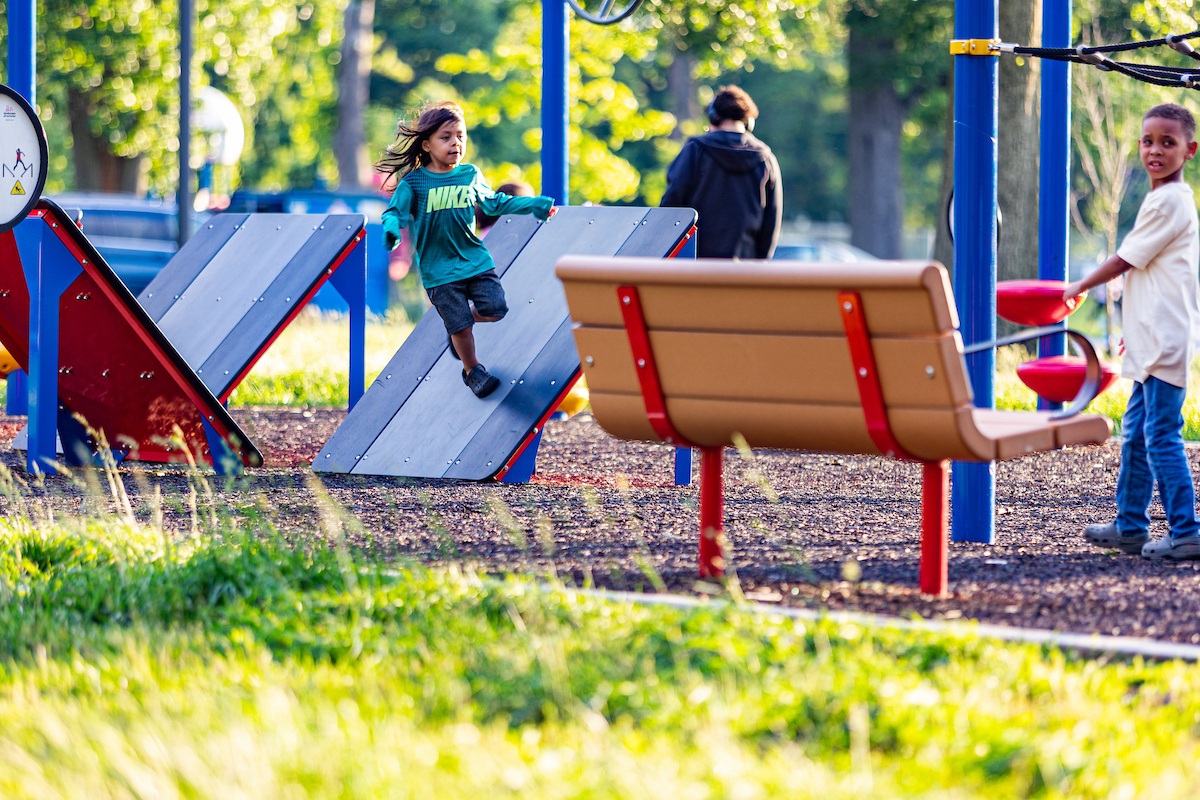 Several children play on outdoor fitness equipment, including slanted platforms for parkour.