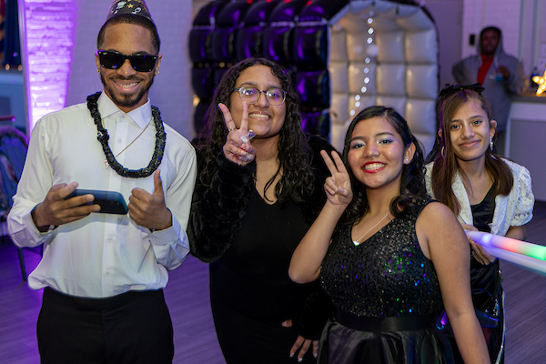 Four smiling teens pose for a photo in formal attire, giving peace signs and thumbs up. One boy wears sunglasses, a novelty party necklace, and a party hat. Behind them, the room is dimly lit with purple LED party lights.