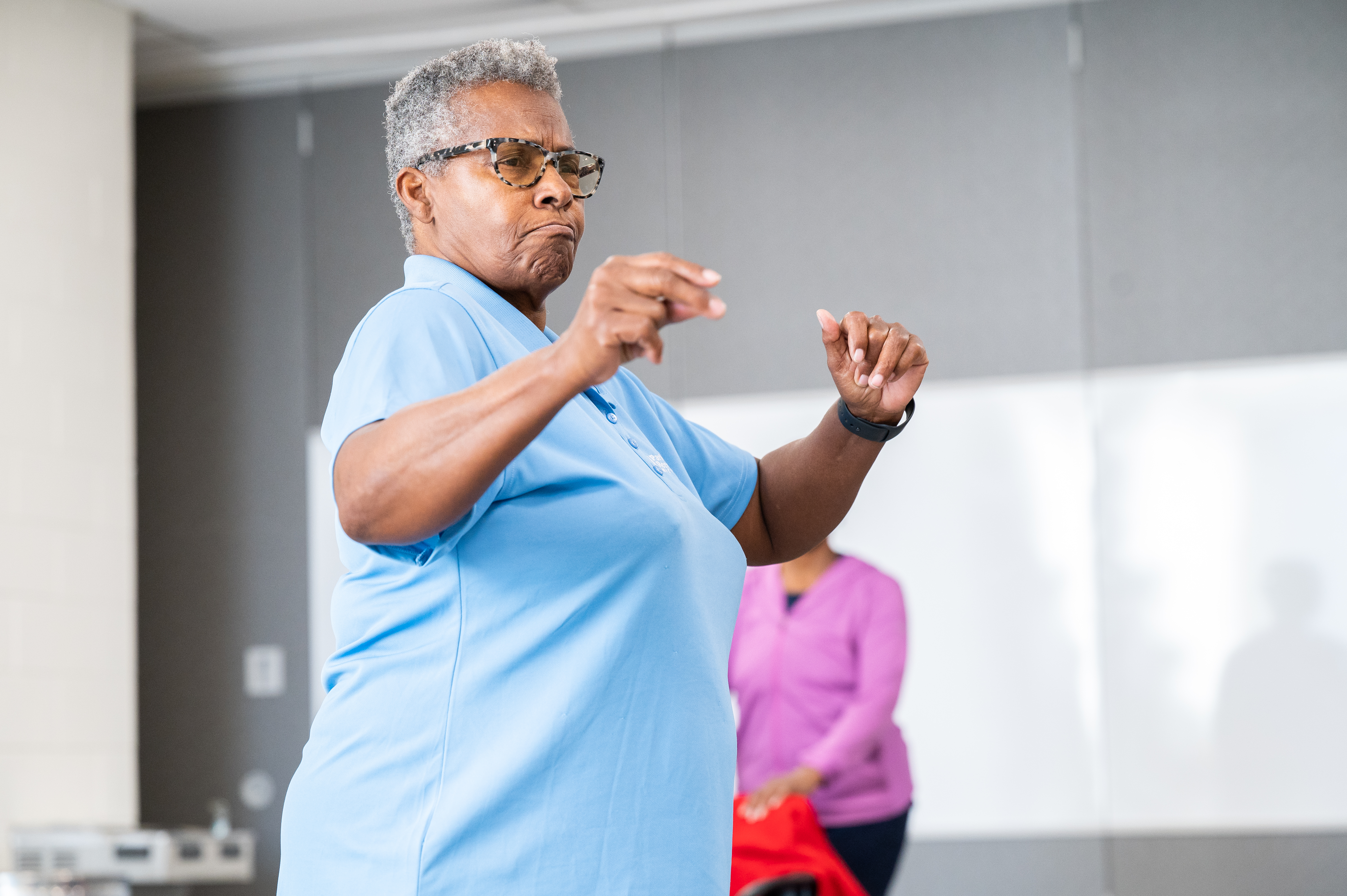 A woman with short gray hair, wearing glasses and a light blue polo shirt, snaps her fingers with a satisfied expression on her face. Behind her is a person in exercise clothing.