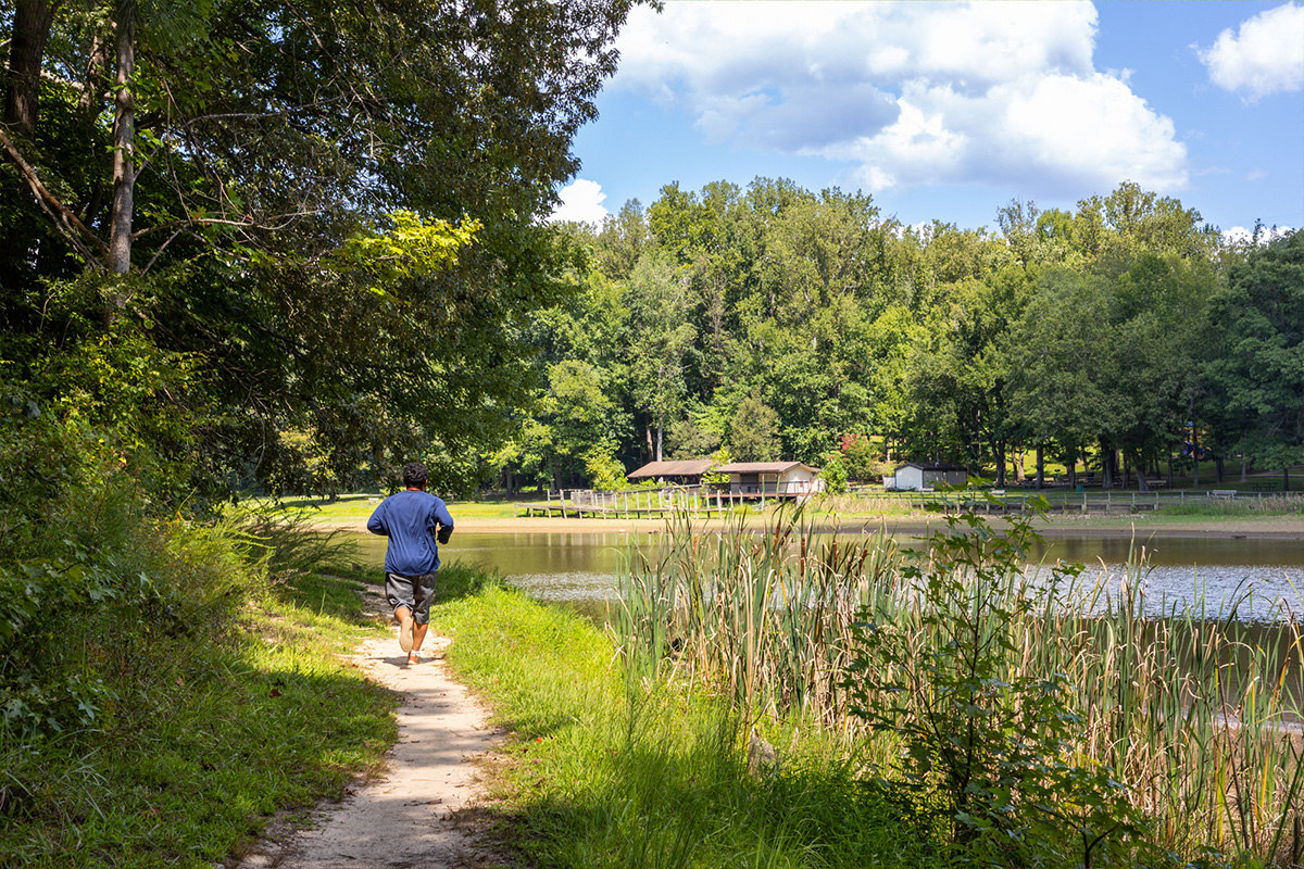 A person jogs on a natural surface trail that runs along the bank of a body of water.
