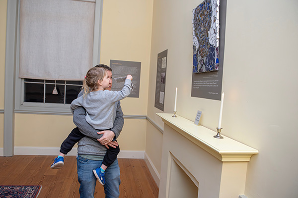 A parent holds a child while they look at an artwork on the wall in a historic room, hanging above a fireplace.