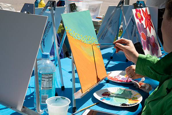 A person paints an abstract artwork on a small canvas resting on a tabletop easel. The table is covered with other paintings-in-progress on easels.