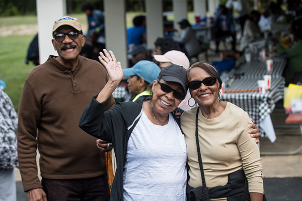 Three seniors smile and wave in front of a large pavilion filled with picnic tables and people.