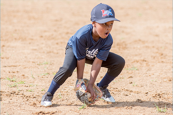 A small boy in a baseball uniform bends down to catch a ball in his glove.
