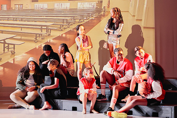 Teen performers in a play sit on bleachers on a stage, some wearing letterman jackets.