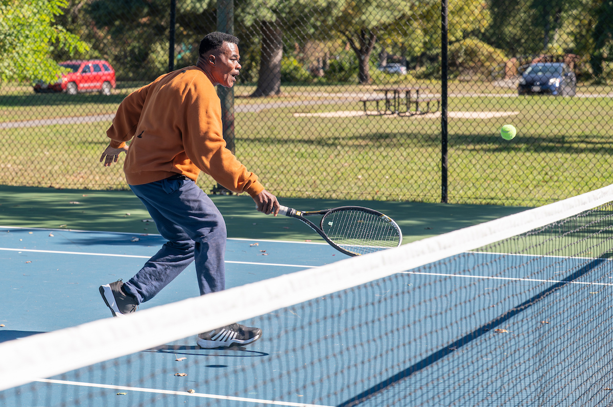 A man plays tennis on an outdoor court. Beyond the fenced court is a field, picnic table, and parking lot.