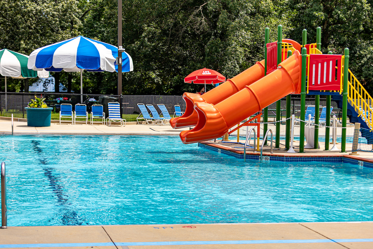 Two side-by-side orange waterslides at a sunny, outdoor pool. There are chairs and shade umbrellas on the far deck.