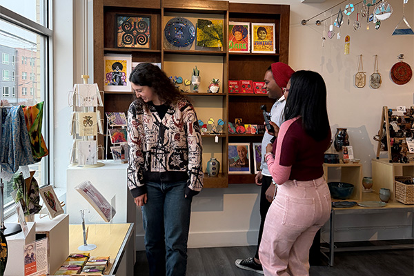 Three smiling customers admire the merchandise in an artisan gift shop. Prints, cards, scarves, sun catchers, and more are displayed near a large window.