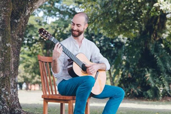 A man plays classical guitar on a wooden chair in the grass, under the shade of a large tree.