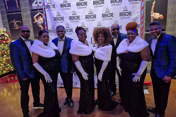 Eight men and women in formal wear pose smiling. The men wear blue brocade jackets and the women wear white fur wraps and long gloves.