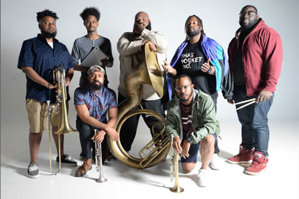 Seven men pose with musical instruments, including trumpets, a trombone, a tuba, and drumsticks.