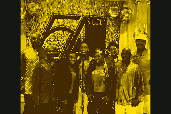 Eight smiling adults pose in front of a backdrop that reads Studio 54.