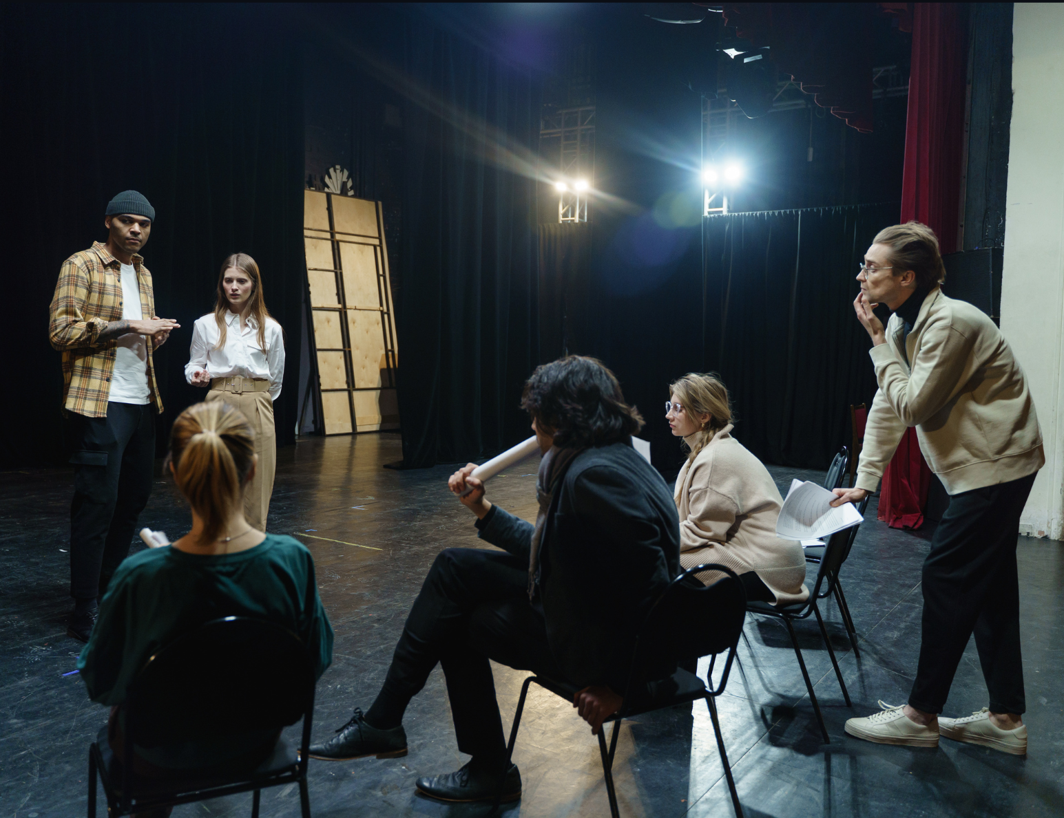 A group of six people during a theater rehearsal. Two actors stand on a stage while four others watch them, holding scripts.