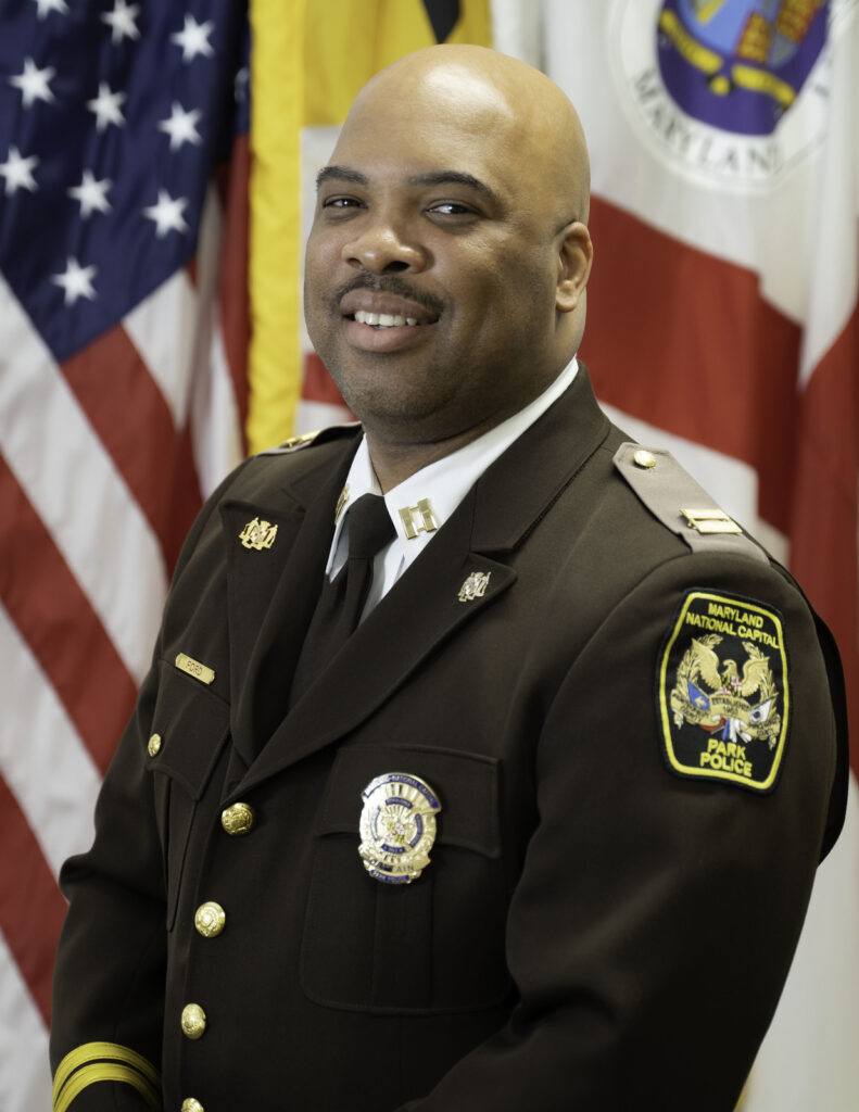 Uniformed park police official in dress uniform, photographed before American, Maryland, and Prince George's County flags.